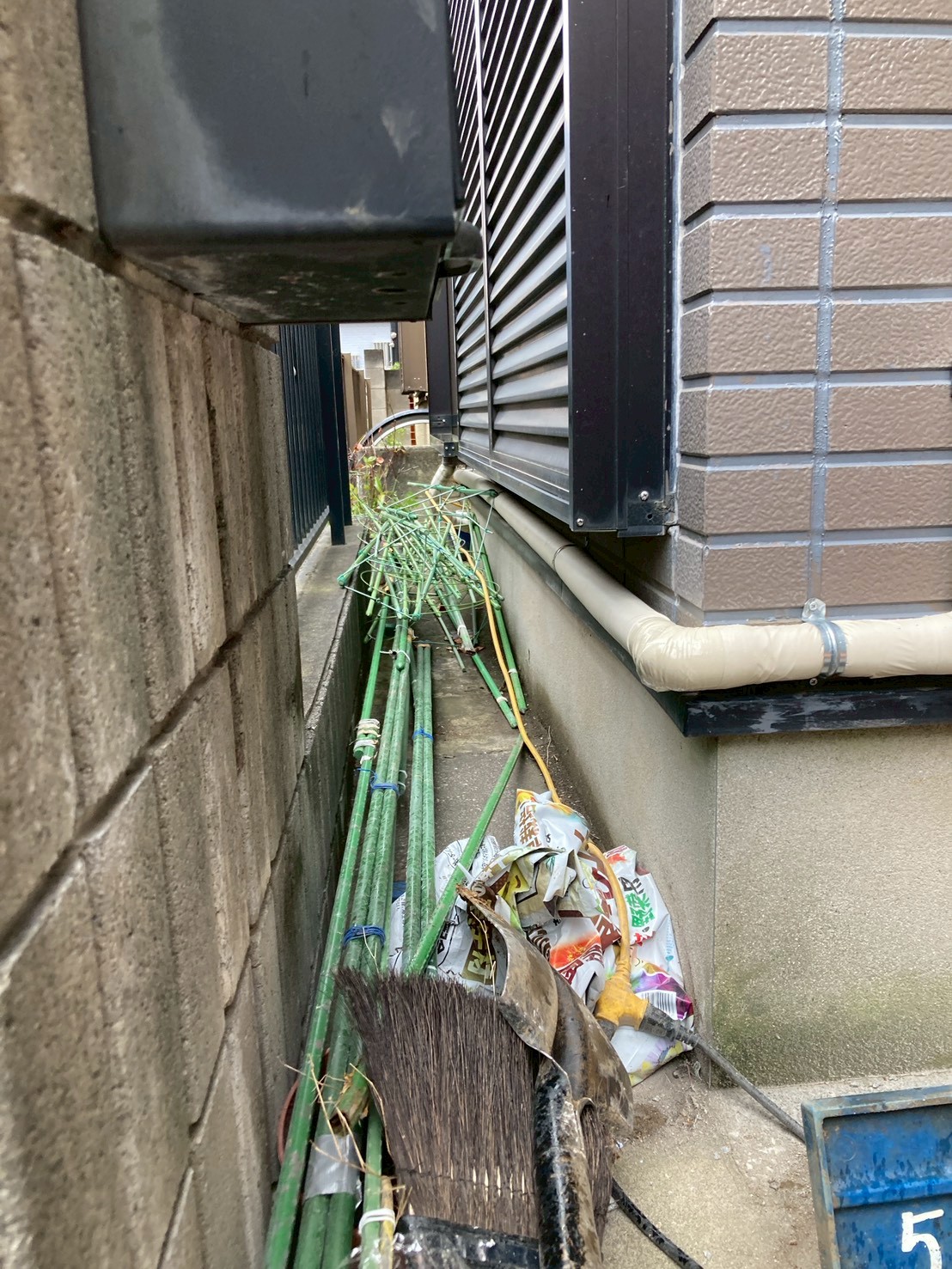 勝手口側通路の現状。古い配管材や不用品が散乱し、地面近くを古い給水管が走っている