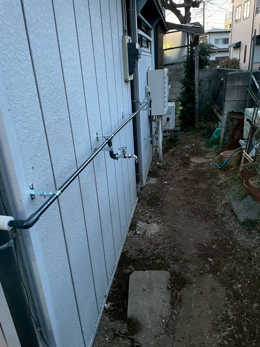 建物正面外壁沿いに新規給水管（黒HIVP）をブラケット固定しながら延伸し先端に新規水栓を設置した状態