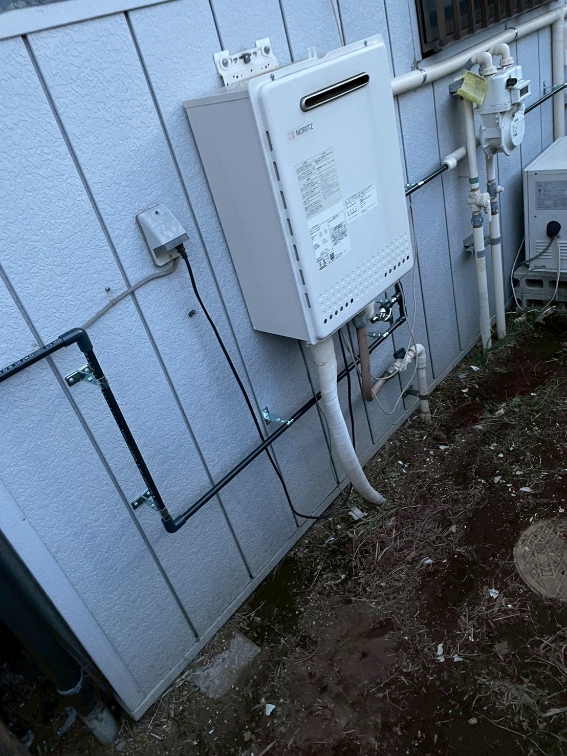 新品NORITZ製給湯器の設置完了。左に新規給水管（黒）が固定され、右にガスメーターまわりの配管が整理されている
