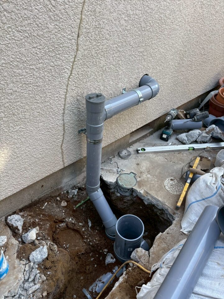 屋外。建物外壁に沿って排水管が立ち上がり、横引き管が壁へ接続される配管全体像