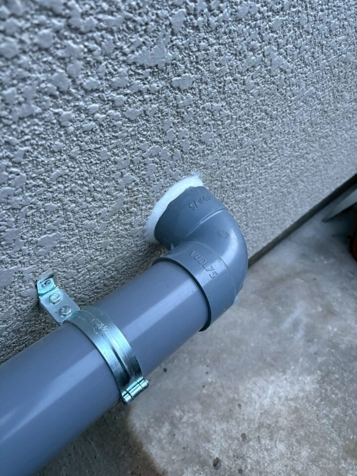 塩ビ管L字継手と外壁貫通部のアップ。バンドとビスで固定されている