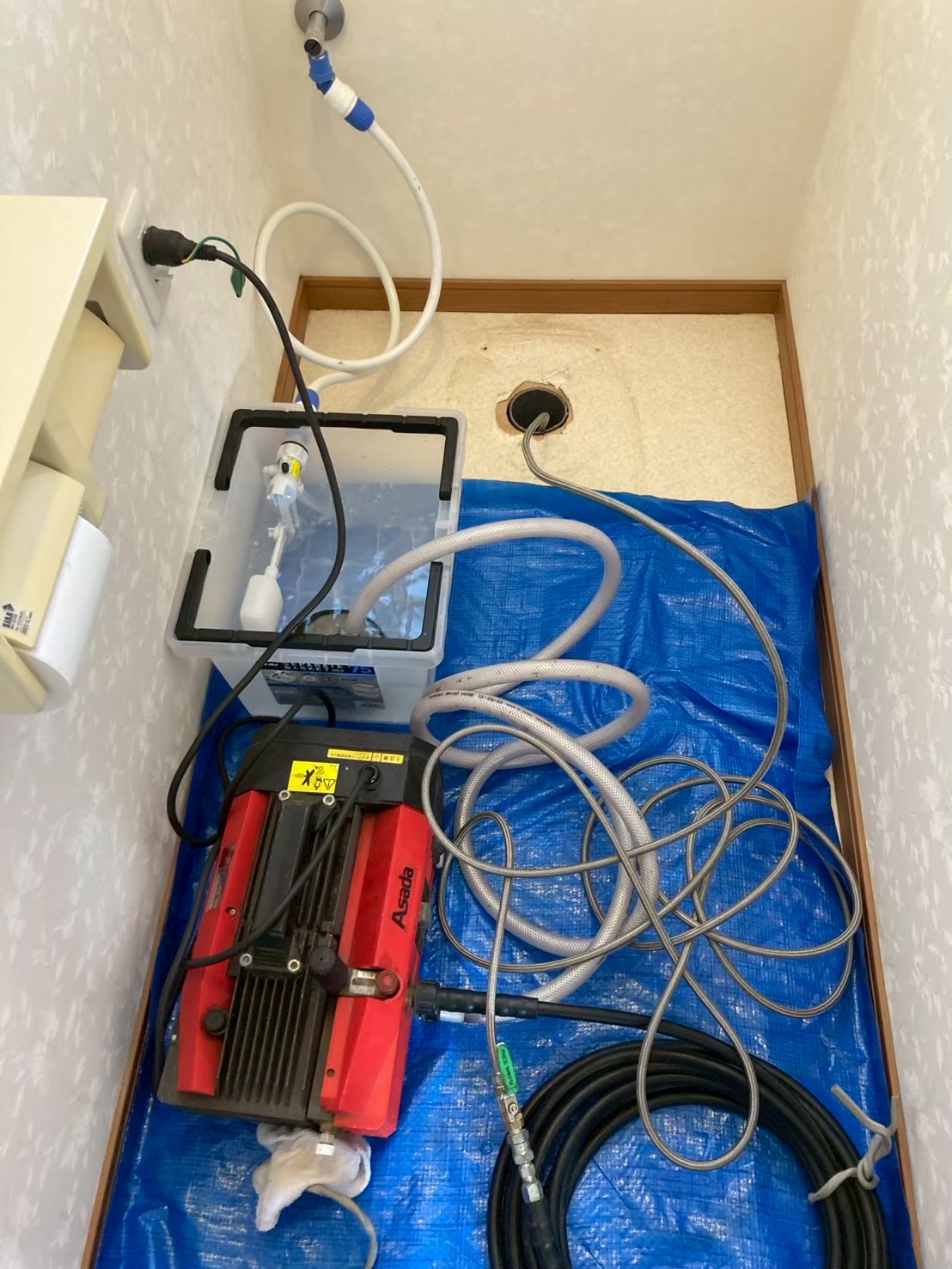 便器を撤去した後のトイレ室。床にブルーシートを敷き、Asada製高圧洗浄機と洗浄ホースを設置して排水管洗浄の準備をしている