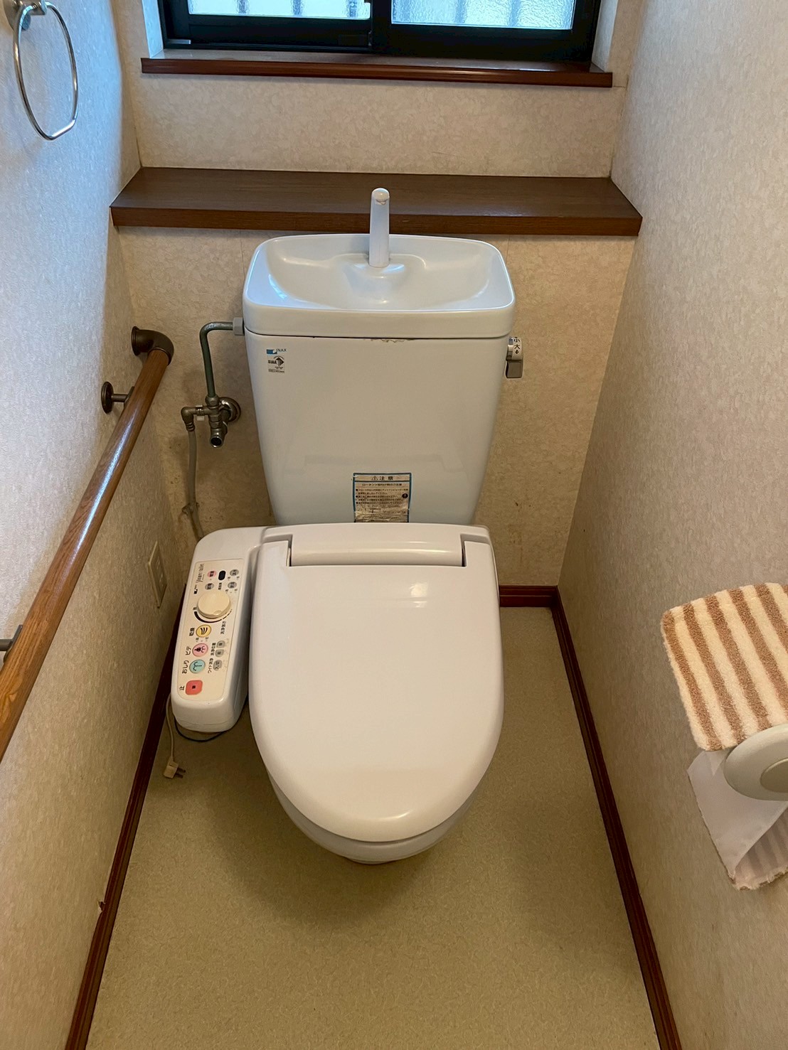 施工前の1Fトイレ。INAX製分離型タンク付きトイレに後付けウォシュレットが設置されている。手すり付き。床には水染みの痕跡がある経年劣化したクッションフロア