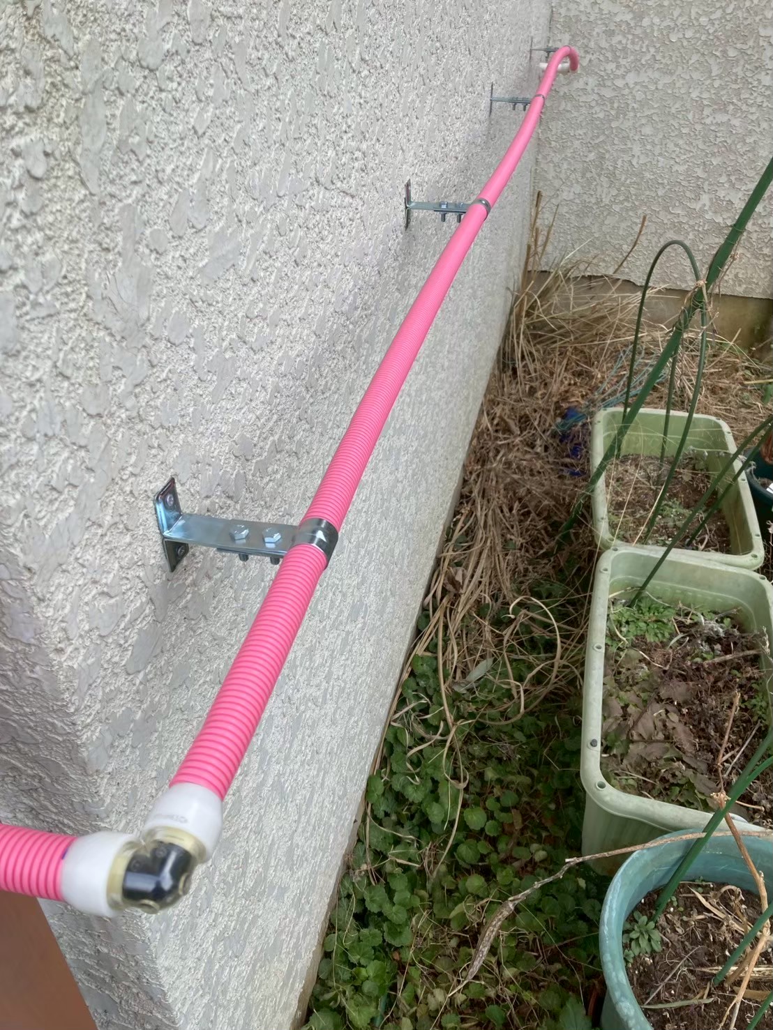 ピンク色のさや管が外壁面に金属ブラケットで固定されて外壁に沿って敷設されている状態