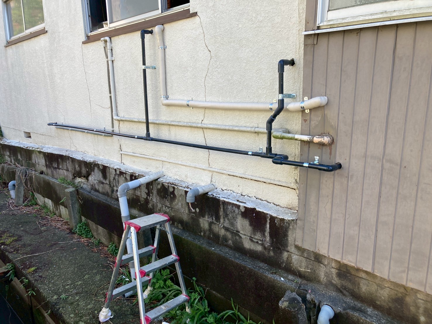 外壁に新旧配管が混在する工事途中の様子。黒い新規配管と既存配管が並行している